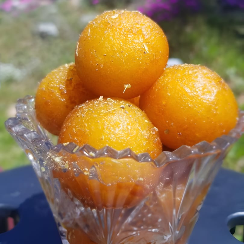 A clear glass bowl filled with golden-orange gulab jamun, sprinkled with white flakes, on a blue table.