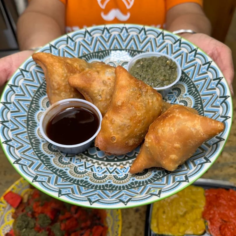 A person holds a colorful plate of golden-brown Cali Tardka samosas with two dipping sauces.