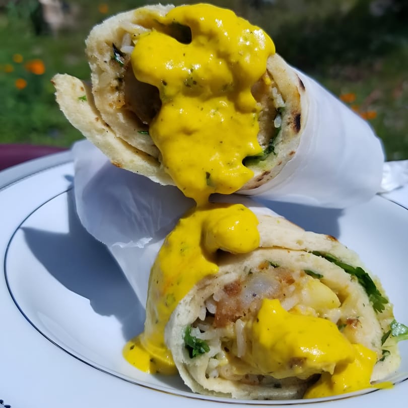 Two Indian takeaway wraps, filled with rice and vegetables, drizzled with yellow sauce on a white plate.
