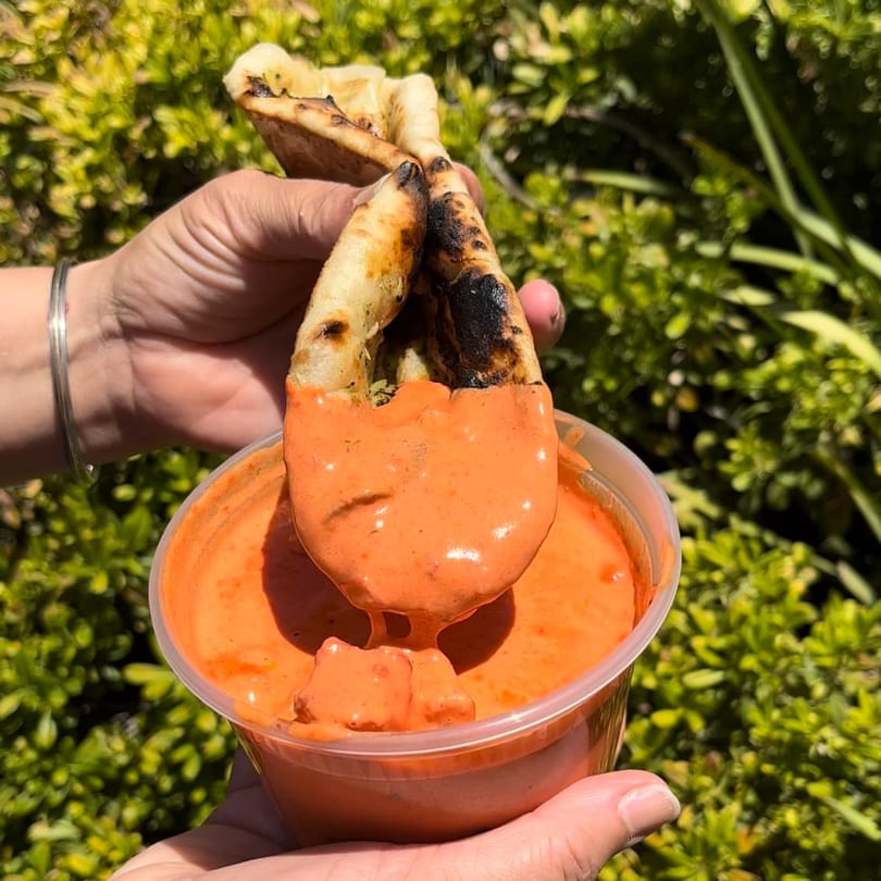 Hands dipping a piece of naan bread into a container of orange Indian curry sauce.