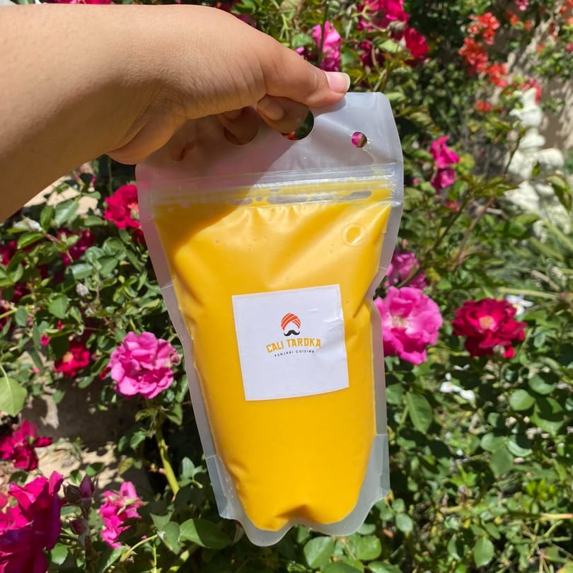 Hand holding a Cali Tadka Indian takeaway pouch filled with yellow curry against a floral background.